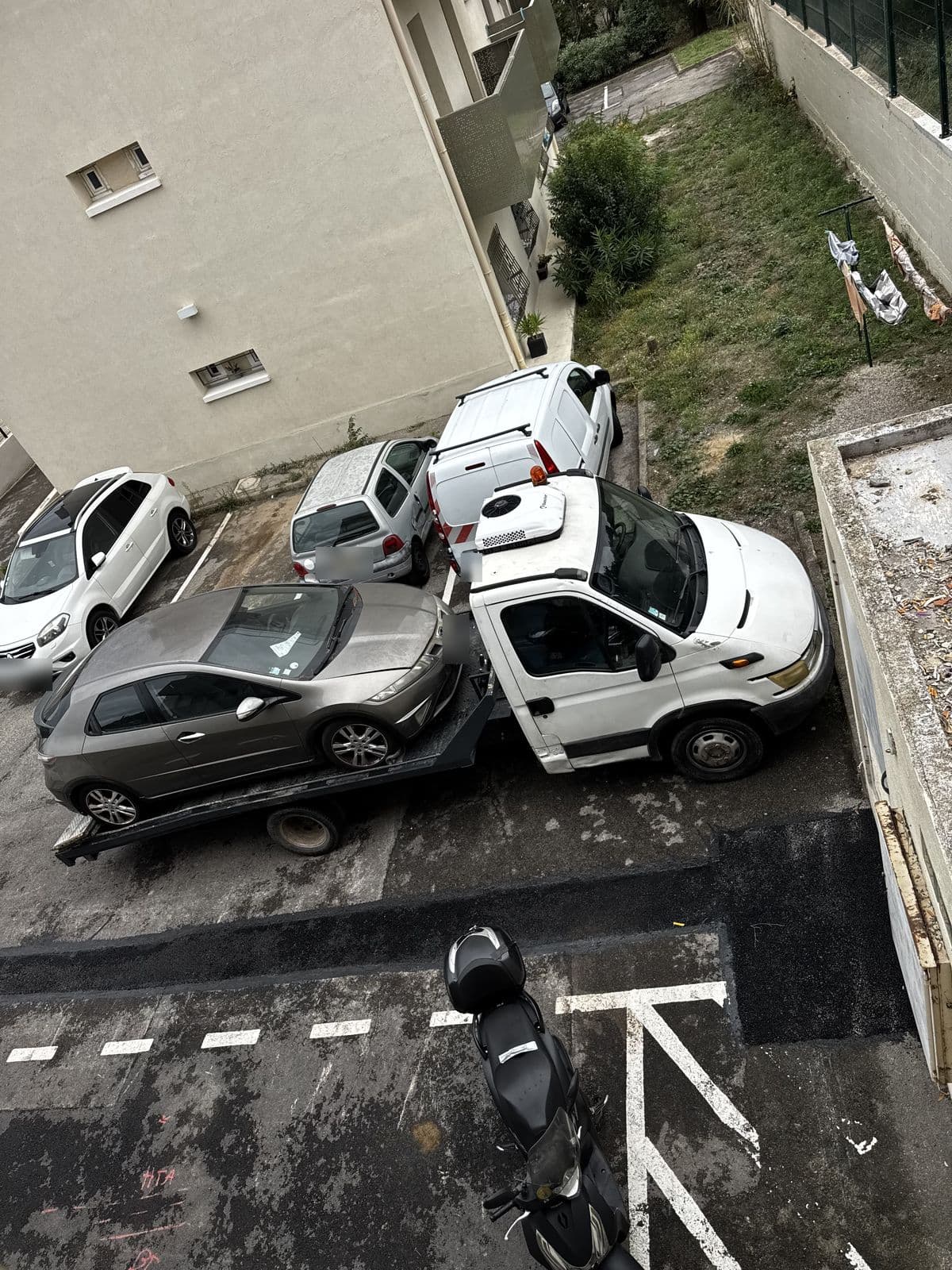 Vue d'en haut du camion plateau chargé d'une berline grise, manœuvrant entre les voitures garées sur le parking d'une résidence