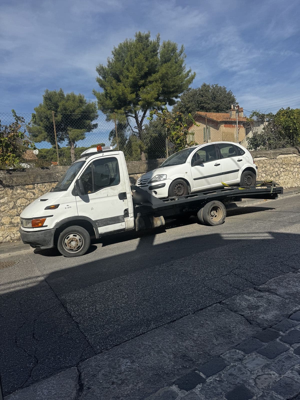 Camion plateau blanc avec une citadine blanche chargée et sanglée, garé le long d'un mur en pierre sèche